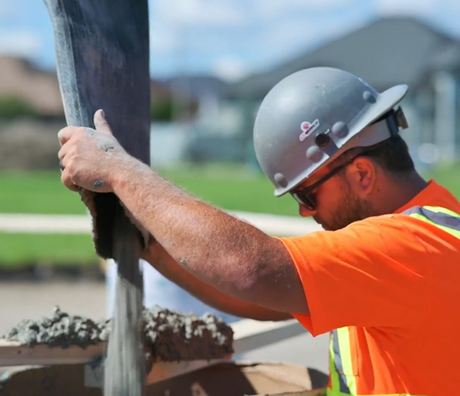 A close-up shot of the Quality Turf crew member wearing a grey hard hat, safety glasses, and an orange high-visibility vest. He is guiding a large black hose as wet concrete is poured into a wooden form. Quality Turf.