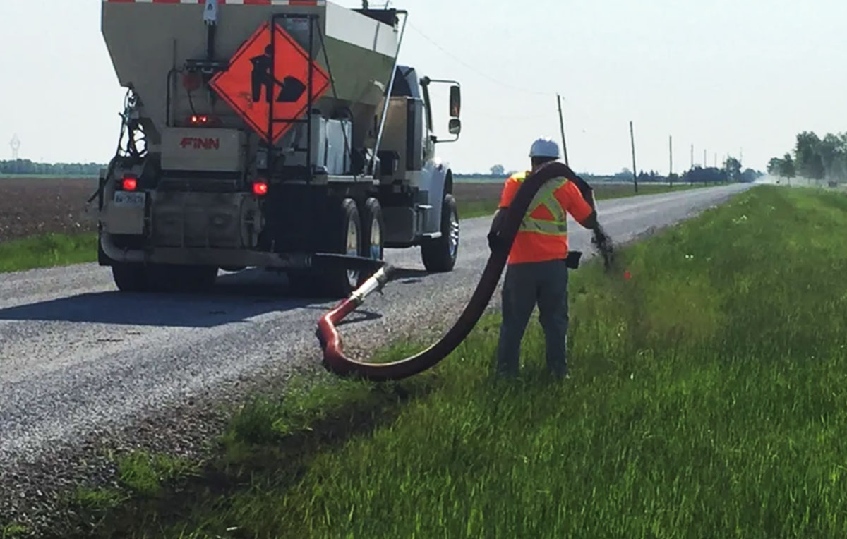 The Quality Turf crew member is using a blower truck hose to spread mulch along the roadside.