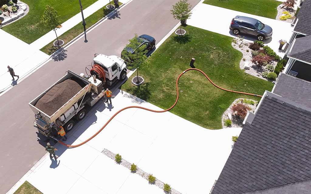 A Quality Turf crew using a specialized bark blower truck and long hose to precisely apply mulch to residential garden beds along a driveway.
