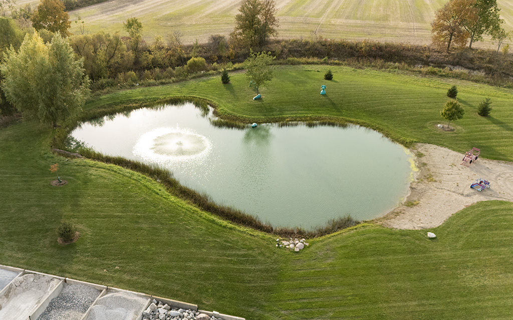 Aerial view of a professionally landscaped backyard featuring a large pond with a center fountain, surrounded by a lush green lawn and manicured trees. Quality Turf.