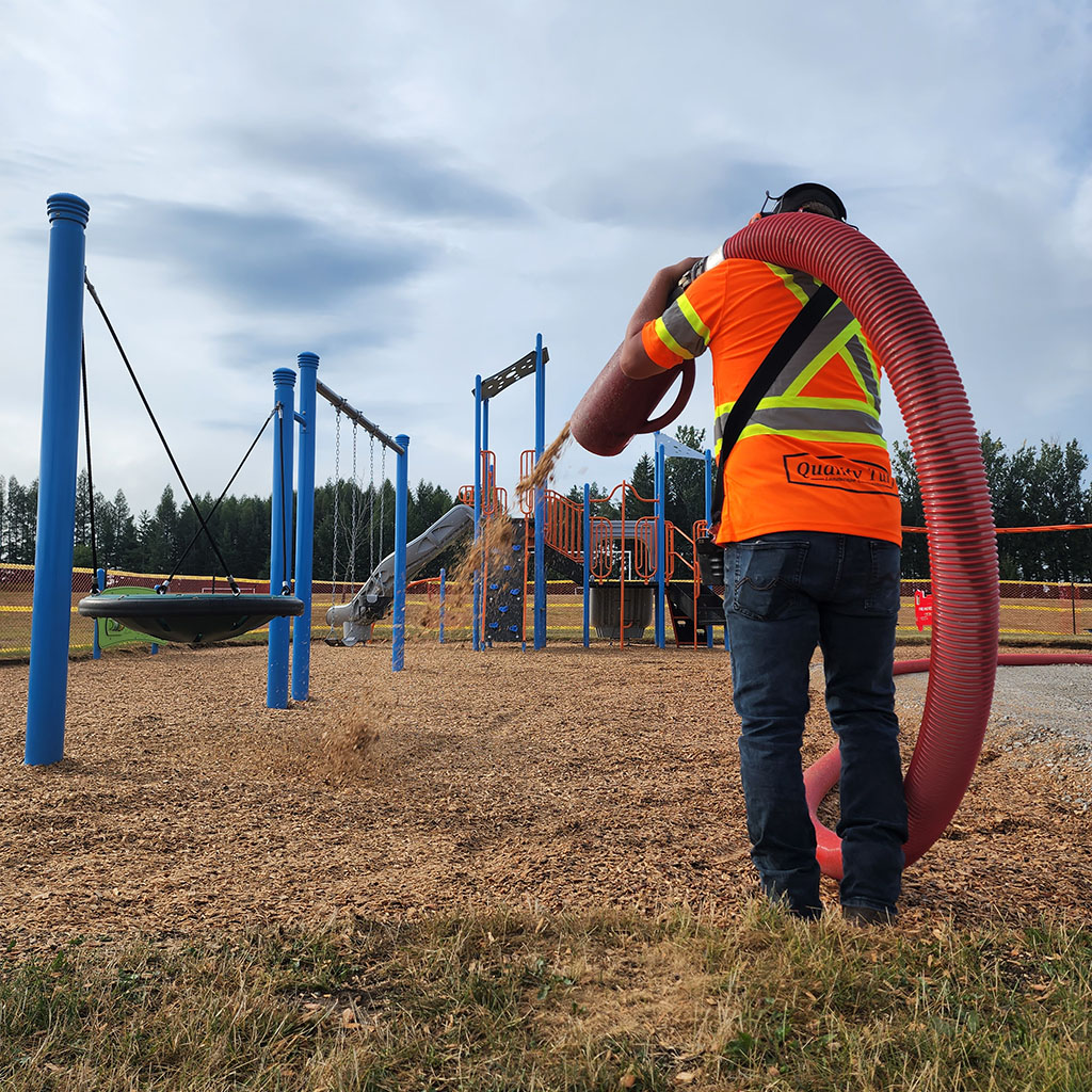 Quality Turf crew member wearing safety gear uses a heavy-duty blower truck hose to efficiently spray fresh wood chips and playground mulch onto a public park play area.