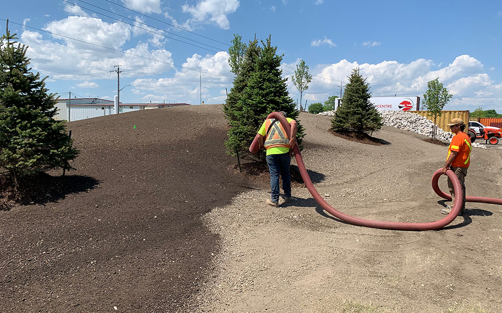 Quality Turf crew members using a high-capacity blower truck hose to evenly distribute dark topsoil and organic compost across a large hillside slope with pine trees.