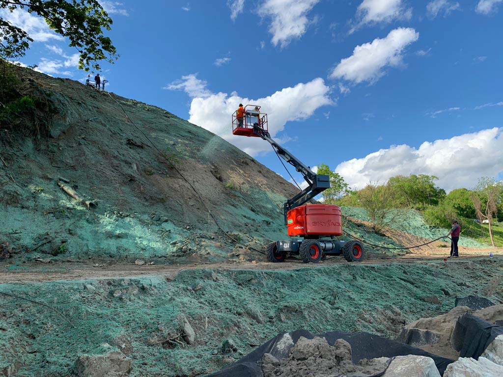 Workers using a boom lift and hose to hydroseed a steep dirt slope.