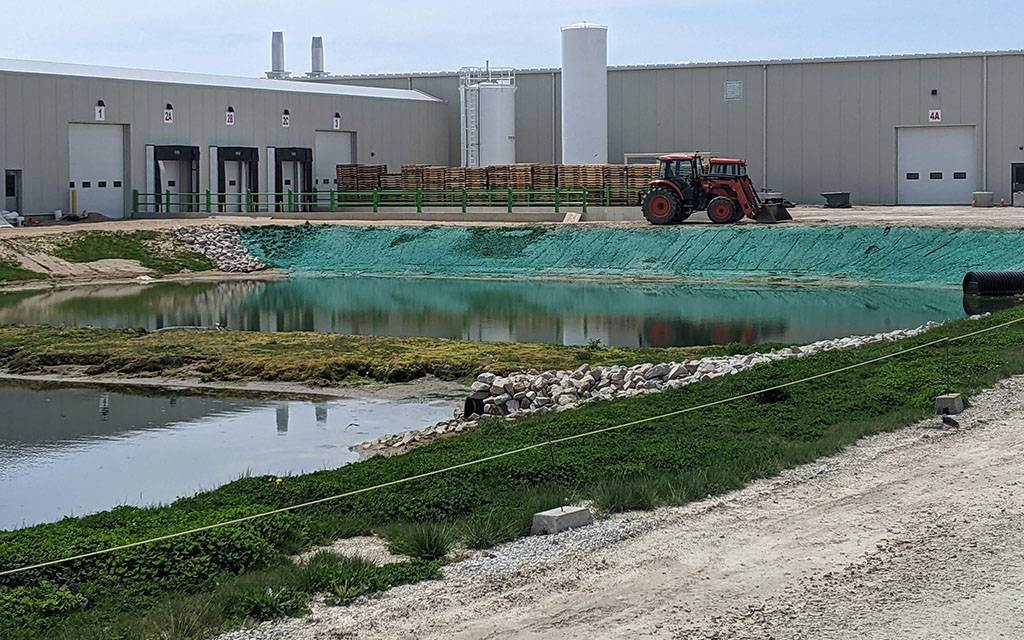 Commercial property shoreline and slope stabilized with green hydroseed slurry near a body of water and industrial building.