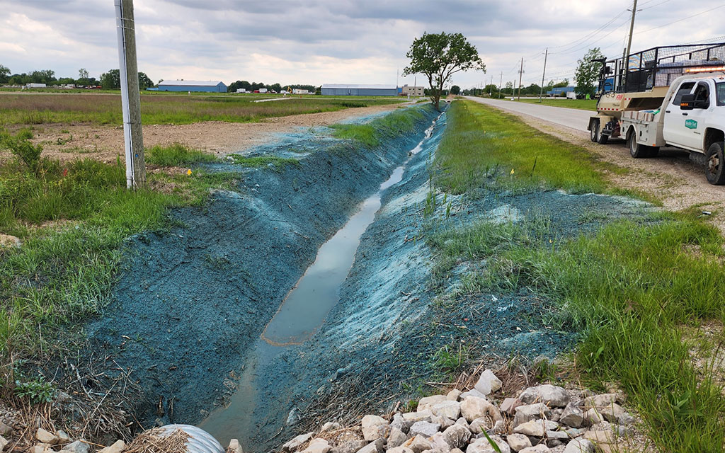 Drainage ditch and roadside slopes freshly sprayed with blue-green hydroseeding mulch for erosion control.