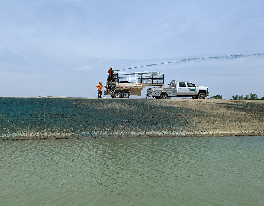 Workers using a professional hydroseeding machine and truck rig to spray seed slurry onto an embankment slope.