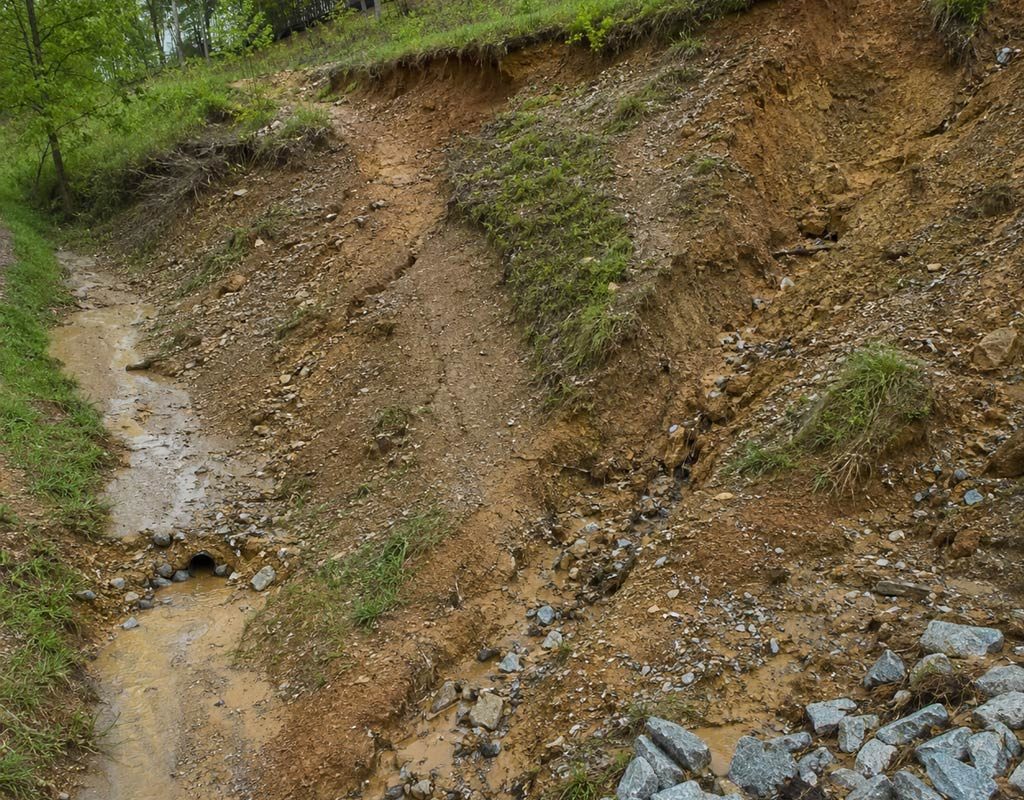 A heavily eroded dirt embankment with visible gullies, runoff water, and rocks at the base.